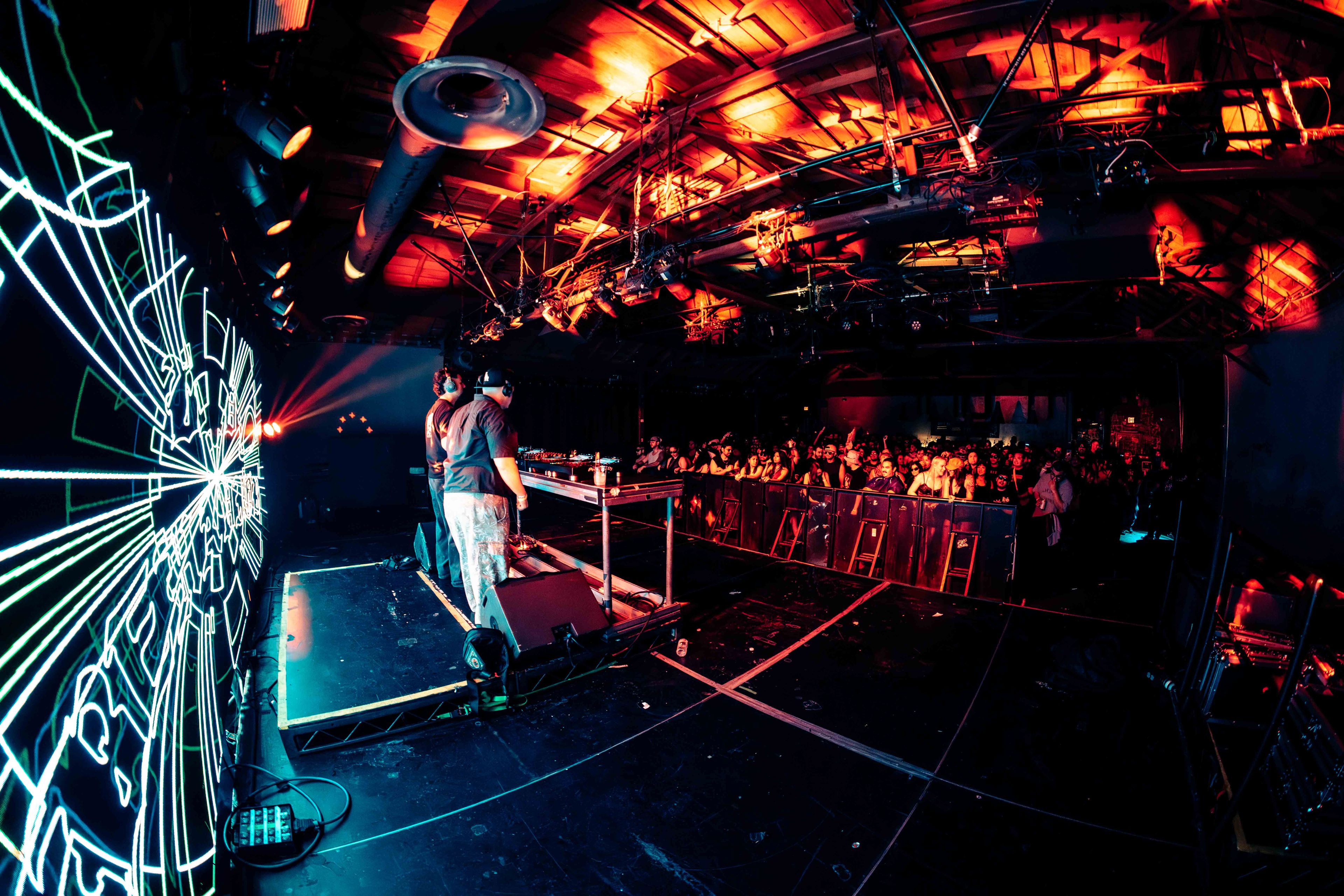 Massive crowd at outdoor festival with stage lights blazing through smoke and fog